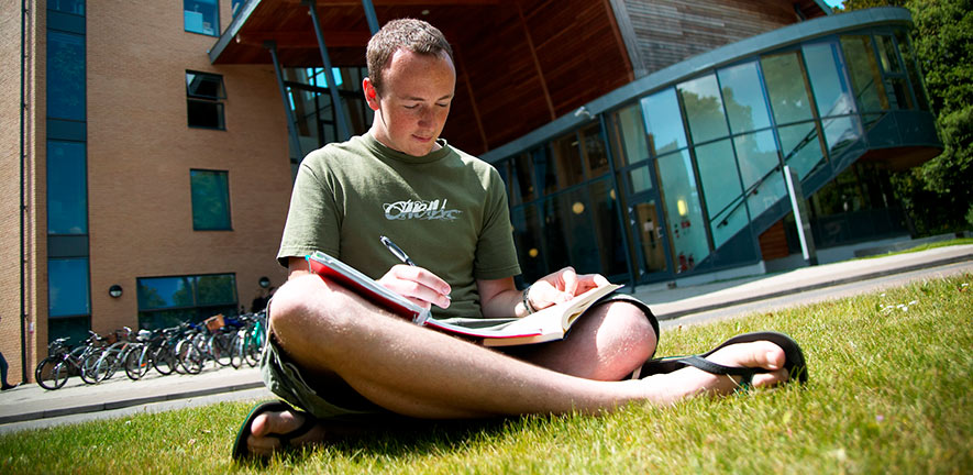 Student reading sits on grass outside of Faculty of Education  