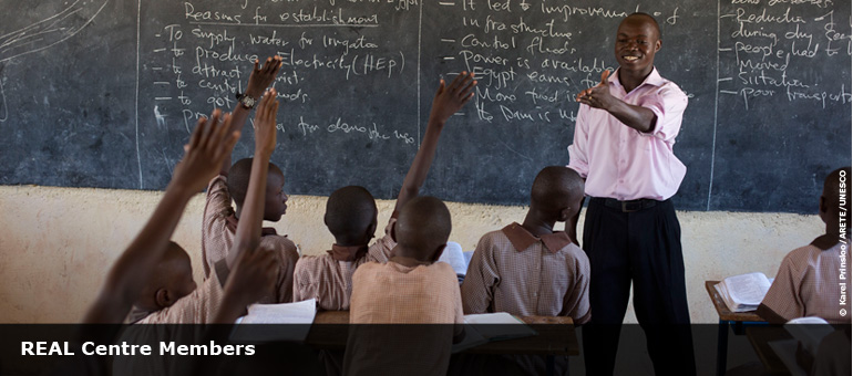 teacher in classroom, in from of chalk board with children holding their hands up to answer a question