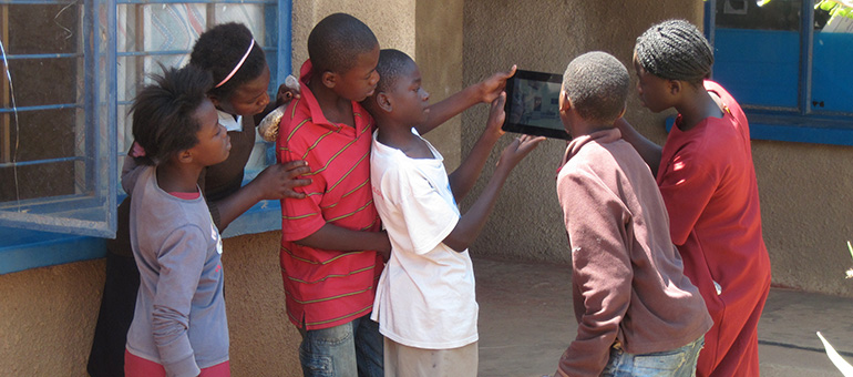 group of happy looking girls and boys stand looking at a digital tablet