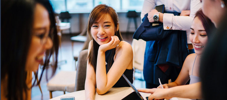 Women professionals sit talking and smiling while working on a tablet