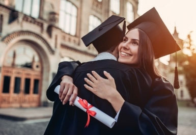 Two students in gowns hug at graduation outside a college building