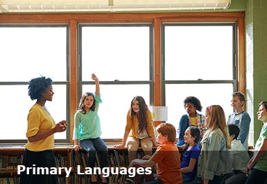 An informal class of school children are focussed on the speaking teacher, one child has a raised hand