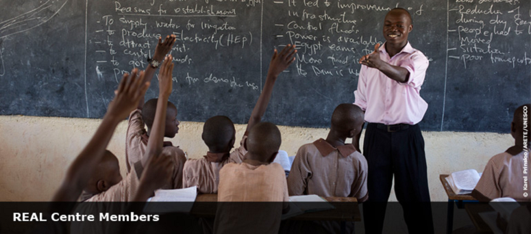 teacher in classroom, in from of chalk board with children holding their hands up to answer a question