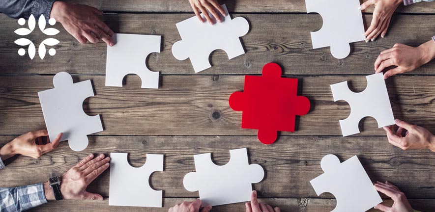 Hands around a table hold large white pieces of a jigsaw puzzle surrounding one large central red piece